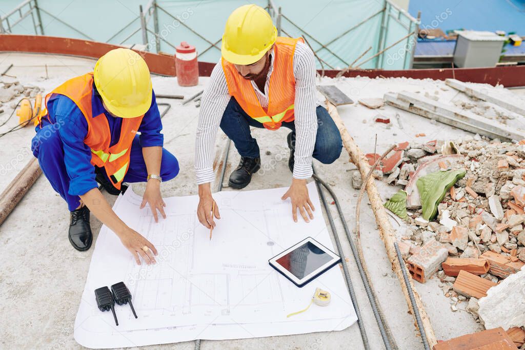 Engineers in hardhats discussing details of house blueprint when working at construction site