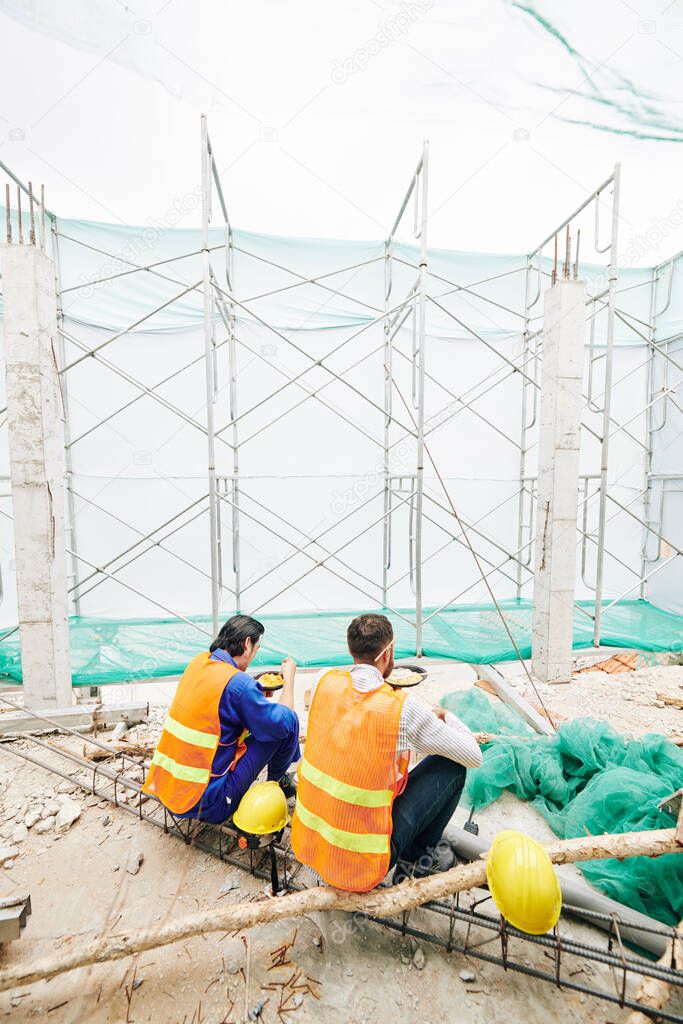 Builders in bright vests eating lunch at construction site, view from the back