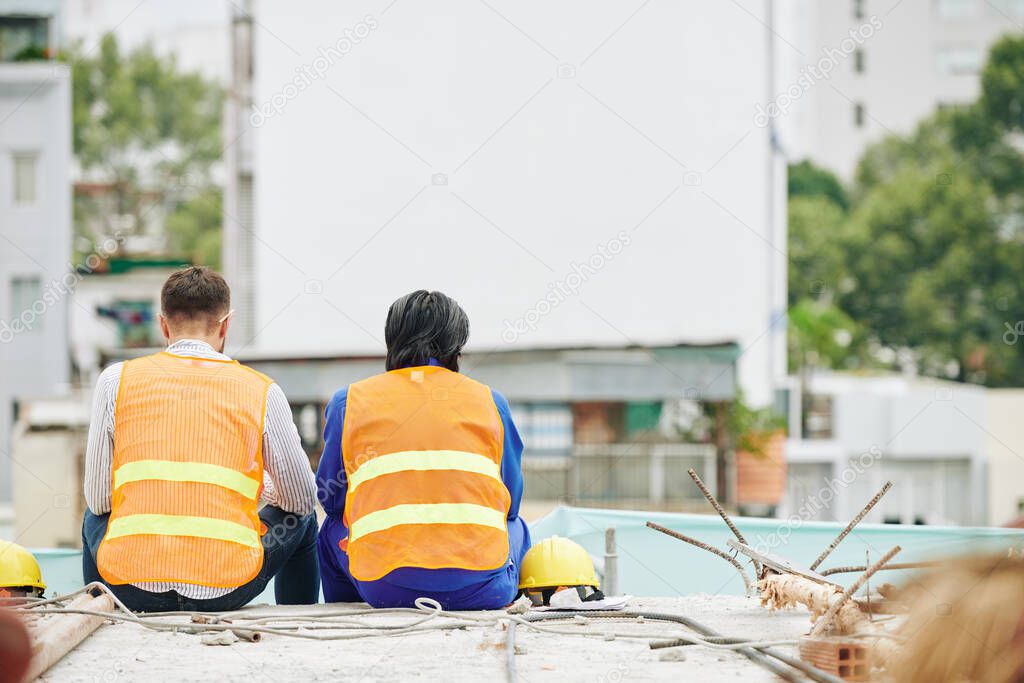 Construction workers in bright orange vests resting after shift and looking at site, view from the back