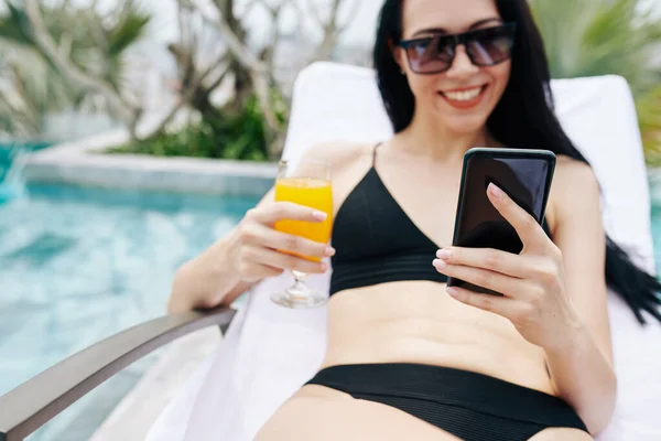 在游泳池边晒日光浴时 带着一杯新鲜橙汁的快乐的年轻女人在智能手机里看短信 — 图库照片