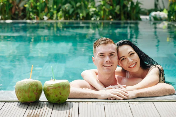 这对年轻漂亮的拥抱情侣靠着椰子鸡尾酒旁边的游泳池边 对着镜头笑着 — 图库照片