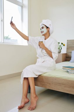 Yüzünde desenlendirici maskeyle yatağının kenarına oturmuş yeşil çay içip selfie çeken mutlu ve güzel Asyalı kadın..