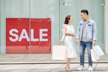 Neşeli genç Asyalı çift büyük mağaza vitrininde satılık afişlerle bekliyor.