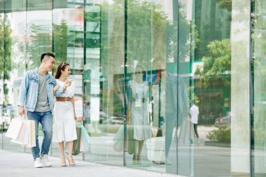 Gülümseyen yakışıklı Asyalı genç adam dükkanın camını işaret ediyor ve kız arkadaşıyla kıyafet konusunu tartışıyor.