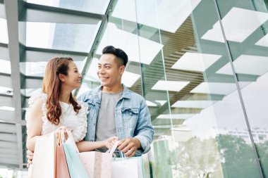 Alışveriş merkezinin önünde duran kağıt torbalı neşeli Asyalı çift.