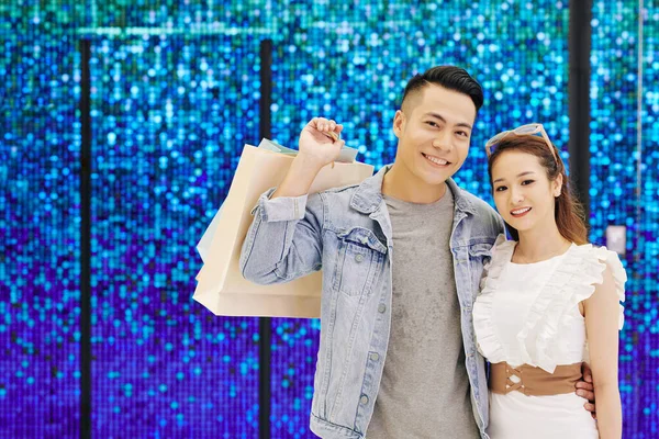 一对快乐的亚洲年轻夫妇站在蓝色闪亮的墙上 提着购物袋 对着相机微笑的画像 — 图库照片
