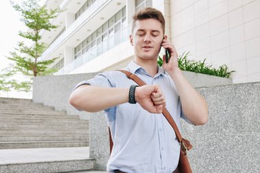 Yakışıklı genç girişimci sokakta yürüyor, telefonla konuşuyor ve kol saatinin saatini kontrol ediyor.