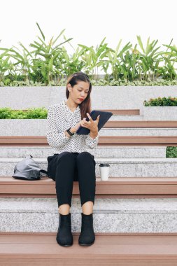 Genç iş kadını açık hava bankında oturuyor ve tablet bilgisayarından mali dökümanları okuyor.