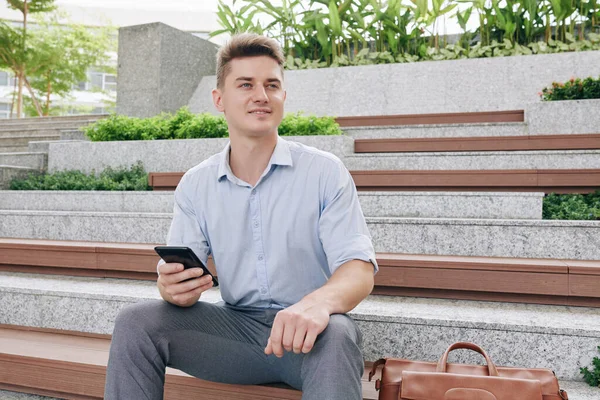 若いです興奮ハンサムなビジネスマンの肖像スマートフォンを手にベンチに座って離れて見て — ストック写真