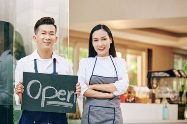Gülümseyen genç Vietnamlı kafe sahipleri açık tabelayla kafe girişinde duruyorlar ve müşterileri içeri buyur ediyorlar.