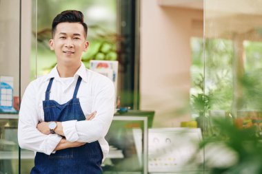 Kendinden emin, kot önlüklü Vietnamlı bir kafenin sahibinin portresi girişte kolları çapraz duruyor.