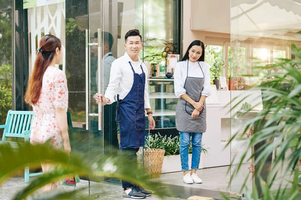 Smiling young waiters standing at cafe entrance doors and inviting customers inside