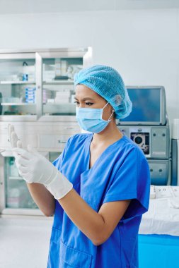 Ameliyat maskesini takıp ameliyat maskesini takan genç, ciddi bir cerrah asistanı sterilize edilmiş ameliyat eldivenlerini giyiyor.