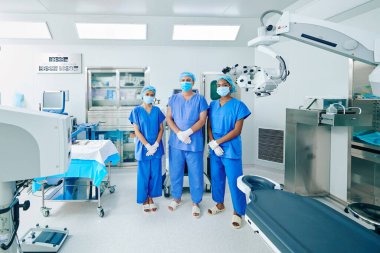 Serious confident surgeons in scrubs and medical masks standing in operating room clipart