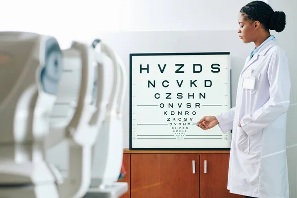 Ciddi Genç Siyah Göz Doktoru Test Panosundaki Mektuplara Işaret Ediyor — Stok fotoğraf