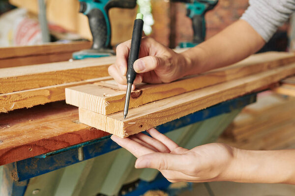 Close-up image of carpenter drawing marks on long wooden plank before cutting it and making furniture unit