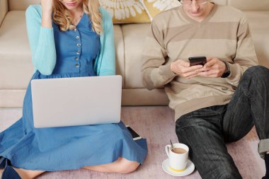 Genç çift oturma odasında yerde oturuyor, kadın dizüstü bilgisayarda çalışıyor erkek kahve içip akıllı telefondan mesajları kontrol ederken.