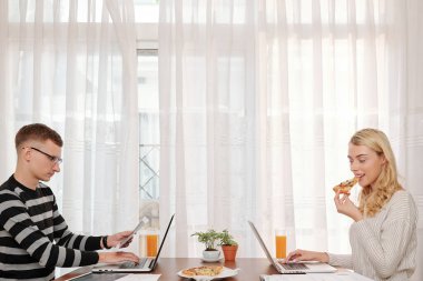 Genç çift evdeki büyük masada oturuyor, pizza yiyor ve dizüstü bilgisayarlarla çalışıyor.