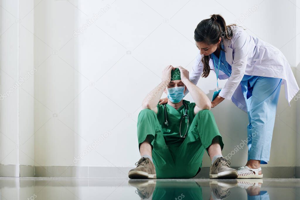 Nurse trying to reassure stressed young surgeon sitting on the floor in clinic corridor after death of patient