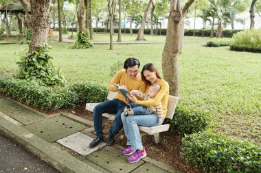 Parkta bankta oturup küçük oğullarına ilginç kitaplar okuyan mutlu ebeveynler.