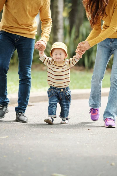 公園を歩くときに彼の両親の手を握って帽子のセーターとジーンズで愛らしい小さなベトナムの少年 — ストック写真
