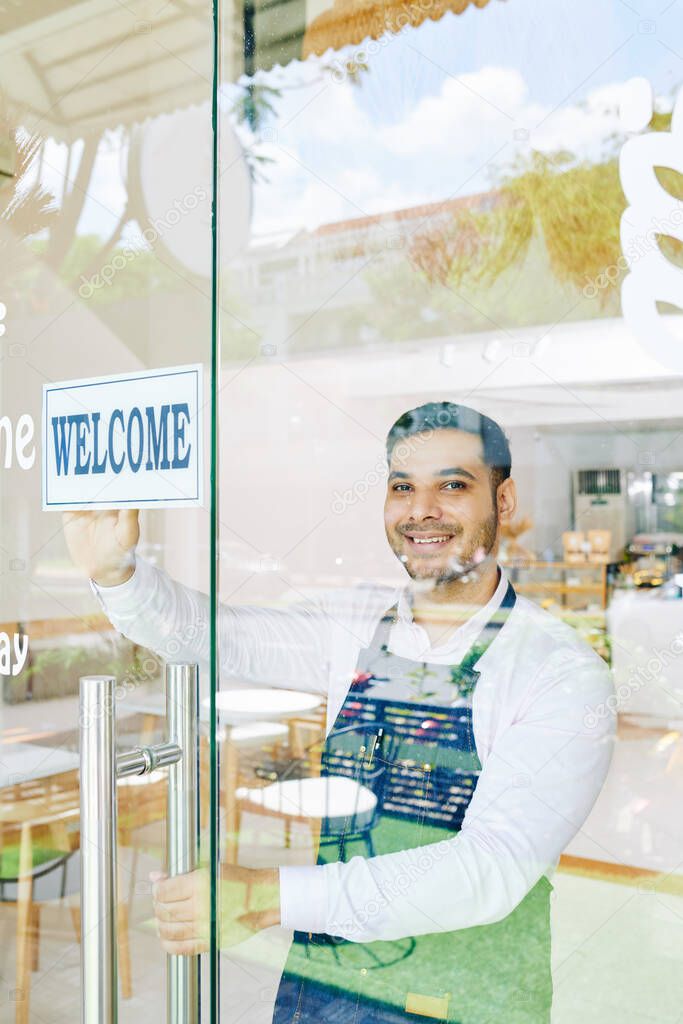 Smiling small bakery owner stocking welcome sign on glass door when opening cafe in the morning