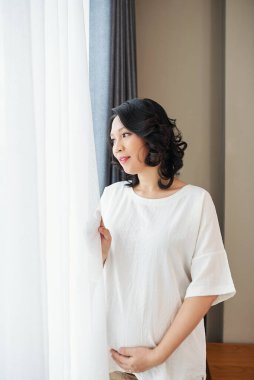 Gülümseyen genç, hamile Vietnamlı bir kadın pencereden bakıp karnına dokunuyor.