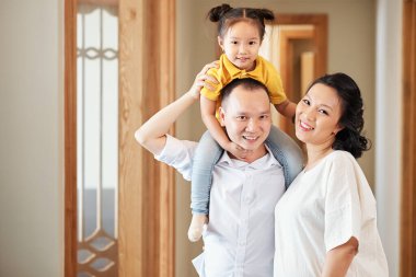 Mutlu gülümseyen Asyalı anne, baba ve küçük kızın portresi daire koridorunda dikilip kameraya bakıyor.