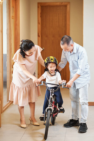 Mother and father helping little daughter in helmet with riding bike in apartment corridor