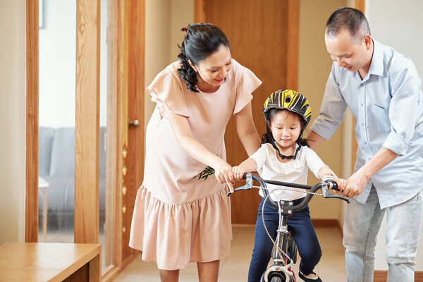 Little Vietnamese girl riding bicycle in apartment corridor with parents helping her to balance on two wheels