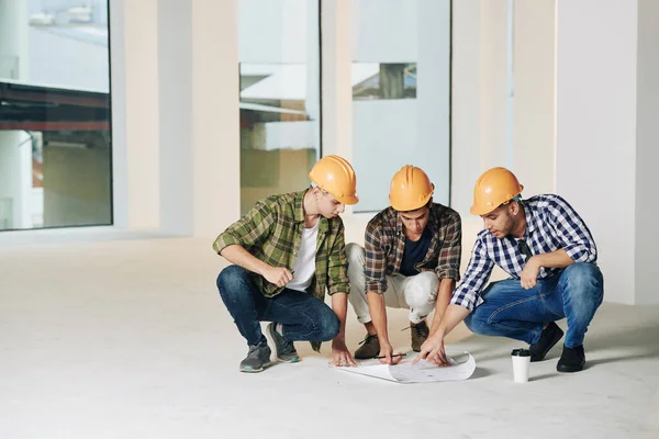 Σοβαρές Νέοι Μηχανικοί Κατασκευών Hardhats Συζητούν Σχέδιο Κατασκευής Στο Πάτωμα — Φωτογραφία Αρχείου