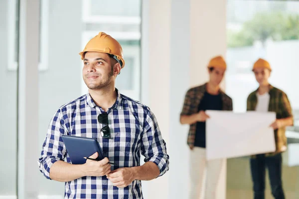 Sonriente Joven Ingeniero Hardhat Celebración Tableta Ordenador Mirando Dentro Casa — Foto de Stock