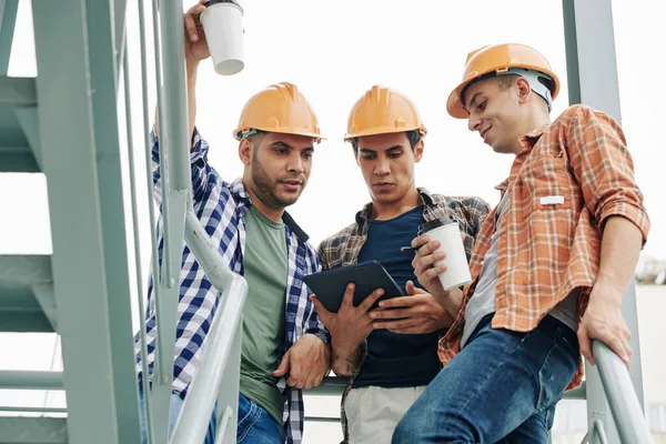 ハード帽子を飲む若い建設労働者は タブレットコンピュータ上の請負業者からコーヒーと電子メールをチェックする — ストック写真
