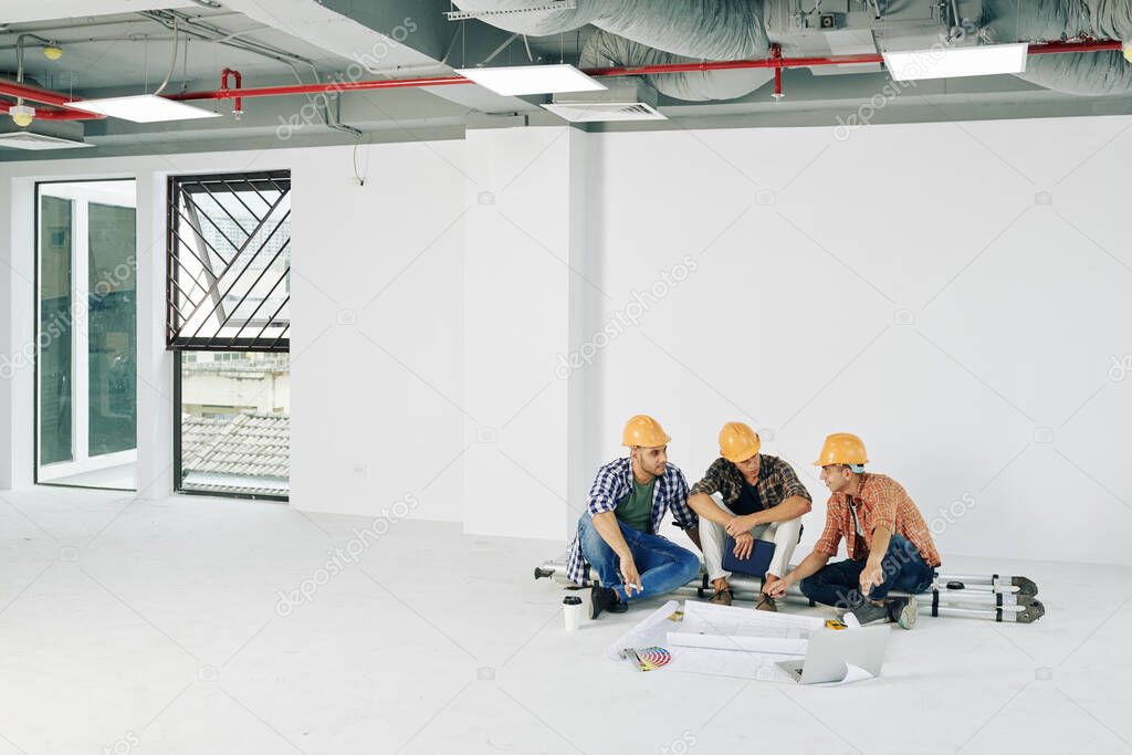 Young construction workers sitting on step ladder with blueprints and color palette in front of them and planning work