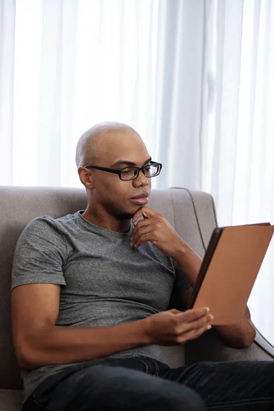 ハンサムな若いです黒男で眼鏡に座ってソファと読書興味深い本上のタブレットコンピュータ — ストック写真