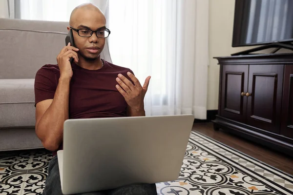 若いアフリカ系アメリカ人のビジネスマンは 彼の膝の上にラップトップで彼のリビングルームに座って 同僚やクライアントと電話で話しています — ストック写真