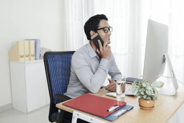 Genç Asyalı girişimci bilgisayar ekranında belge okurken meslektaşıyla telefonda konuşuyor