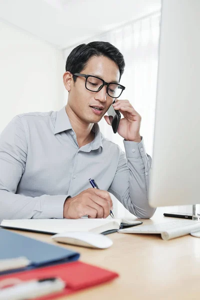 年轻的亚洲企业家通过电话与商业伙伴交谈 并在计划人员中书写 — 图库照片