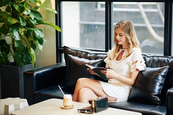 Çekici genç sarışın kadın kanepede dinleniyor, buzlu kahve içiyor ve roman okuyor.