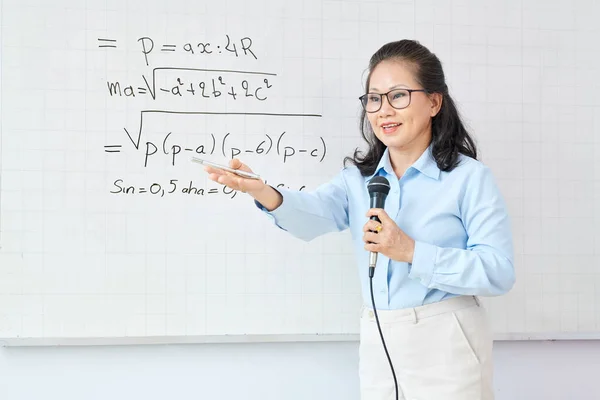 白いボード上の方程式を解決するために学生を求めるときにマイクで話す成熟したアジアの女性数学教師 — ストック写真