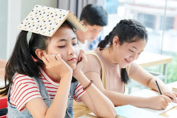 Unatkozó Tinédzser Lány Iskolaasztalnál Nyitott Könyvvel Fején Miközben Mosolygó Osztálytársa — Stock Fotó