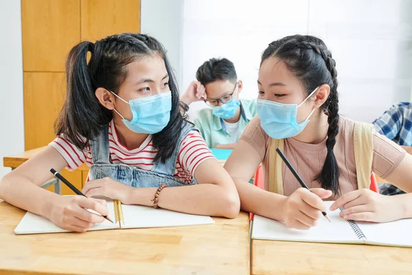 Vietnamese schoolgirls in medical masks sitting at desk in class and discussing difficult task