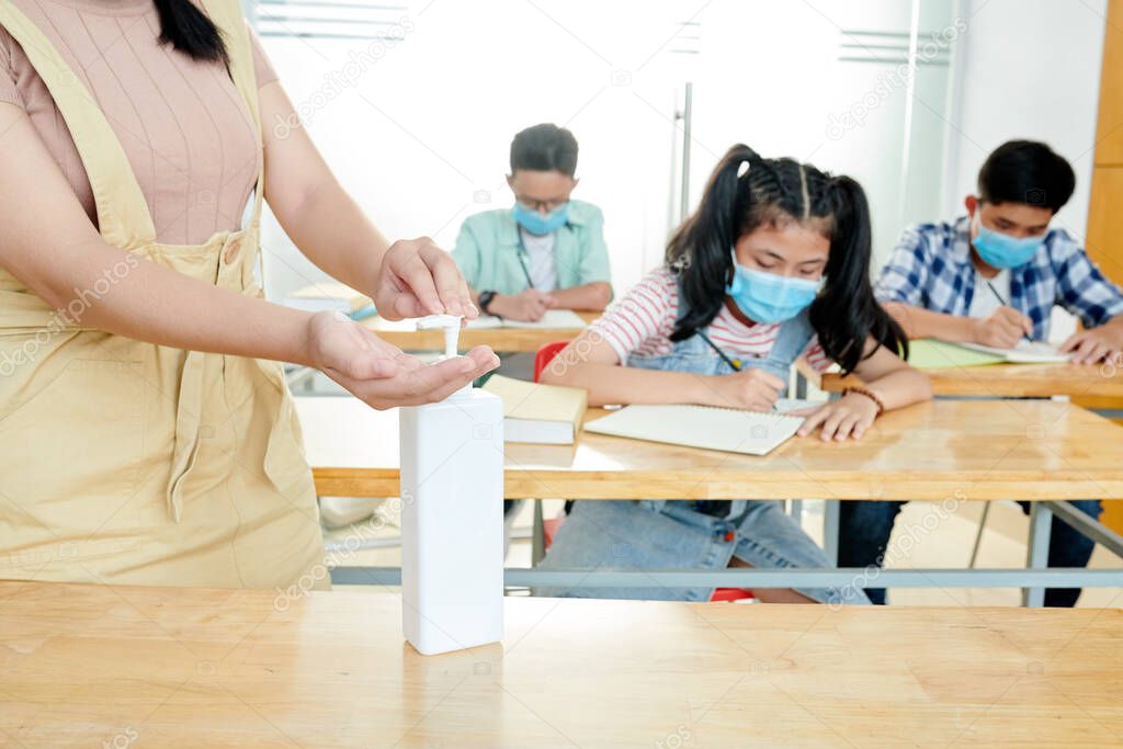 Teacher applying disinfecting gel on hands when students in medical masks sitting at desks and writing in copybooks