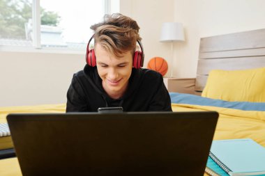 Yatakta dizüstü bilgisayarını açıp akıllı telefonundaki mesajları kontrol eden gülümseyen genç çocuk.