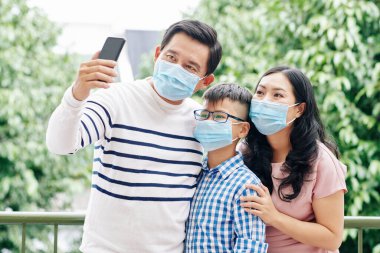 Koronavirüs salgınından dolayı maske takan olgun bir Asyalı. Karısı ve çocuğuyla selfie çekiyor.