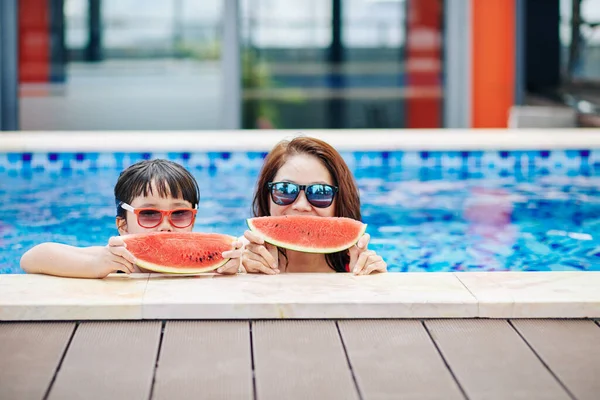 快乐的母亲和戴着太阳镜的小女儿站在游泳池里 吃着新鲜的西瓜 — 图库照片