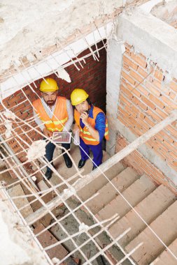 Tablet bilgisayarlı inşaatçılar inşaat halindeki merdivenlerde duruyorlar, telsizle konuşuyorlar ve yukarı bakıyorlar.