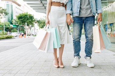 Moda mağazasının önünde alışveriş torbalarıyla el ele tutuşan genç adam ve kadının kesilmiş görüntüsü.