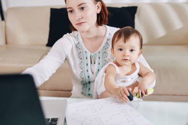 Annesinin kucağında oturan, dizüstü bilgisayarda çalışan ve mali belgeleri kontrol eden tatlı küçük bir kız.