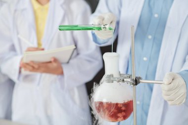 Kauçuk eldivenli öğrencinin tüten sıvıyla deney kabına yeşil reaktör dökerken yakın plan görüntüsü. Sınıf arkadaşı deney sonuçlarını yazıyor.